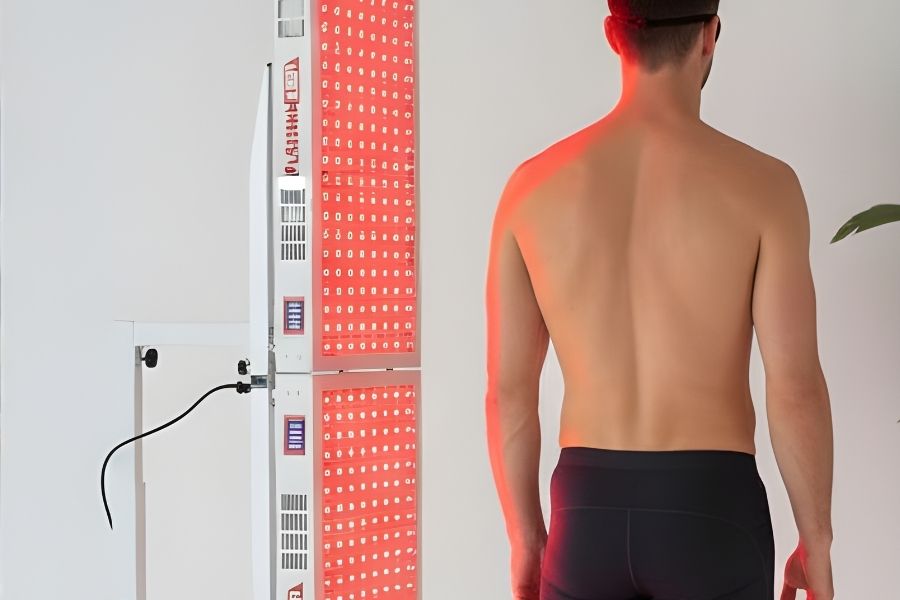 Shirtless man standing before LED red light therapy panel for full-body recovery and pain relief at home