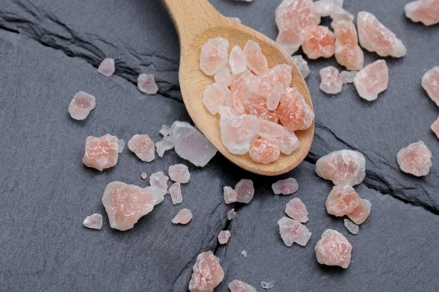 Coarse Himalayan pink salt crystals on wooden spoon scattered on slate background, gourmet seasoning.