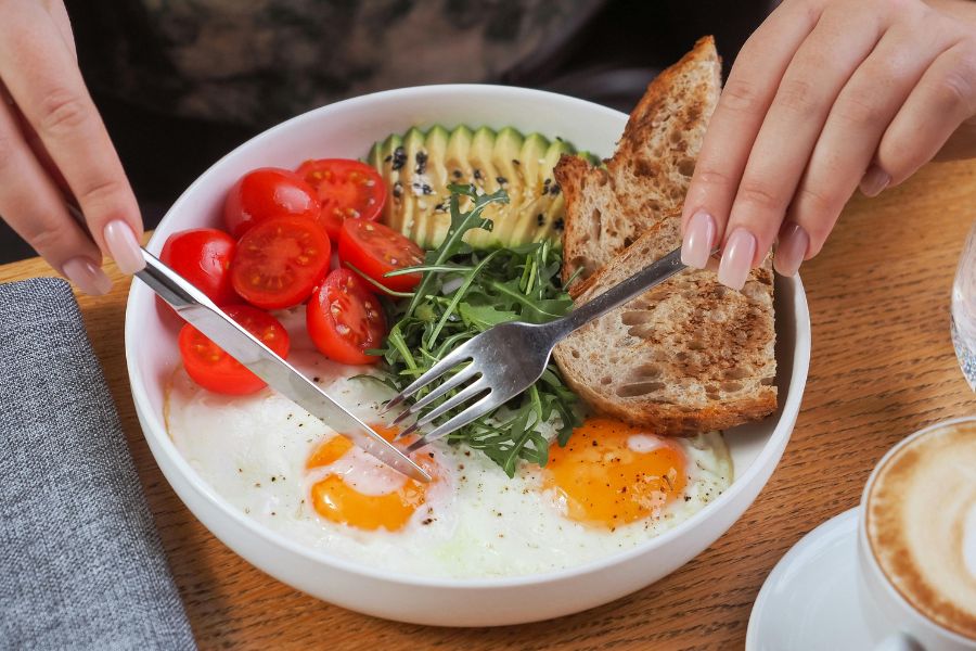 Healthy breakfast bowl with sunny-side eggs, avocado, cherry tomatoes, arugula and sourdough toast, with cappuccino.