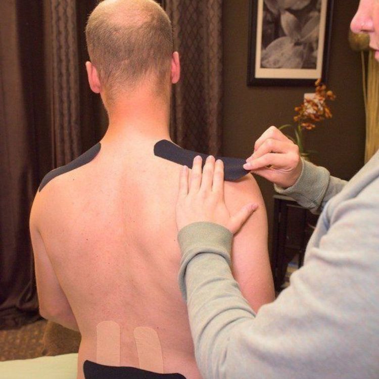 Physical therapist applying kinesiology tape to patient's shoulders and upper back for pain relief and posture support.